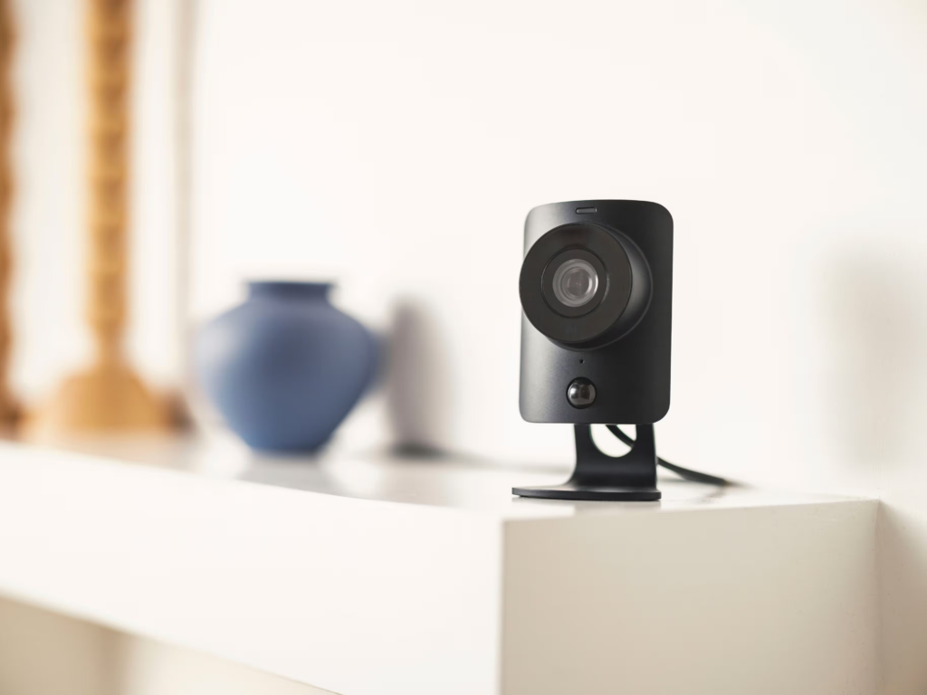 A modern SimpliSafe continuous recording camera displayed on a white shelf, elegantly positioned alongside a blue ceramic vase. The camera features a sleek design with a prominent lens, blending seamlessly into a contemporary home environment.