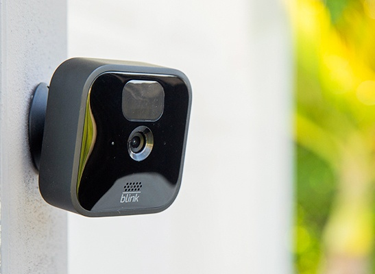 A Blink outdoor camera, a sleek and compact Motion Detector Home Security Cameras, is mounted on a vertical post. The camera features a glossy black finish, a prominent lens at the center, and a motion sensor above it.