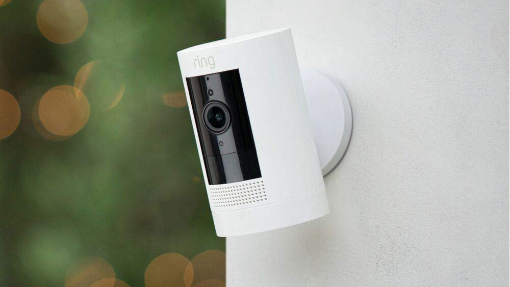 A Ring Stick Up Cam Battery, a versatile Motion Detector Home Security Cameras, is mounted on a wall. The camera features a cylindrical white body with a black front panel housing the lens and motion sensor. 